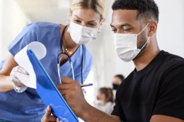 一名男性病人在写字板上签署文件，写字板由一名戴着手术服和听诊器的女医务人员拿着。