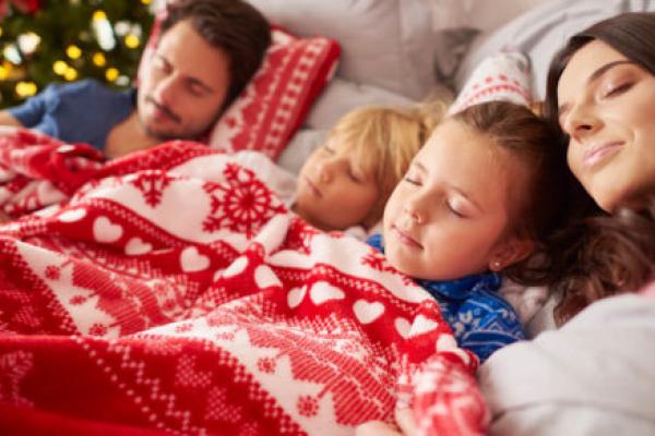 一个男人，女人和两个幼儿躺下，睡觉，覆盖着一个假期主题的毯子。一棵点燃的圣诞树在后台。