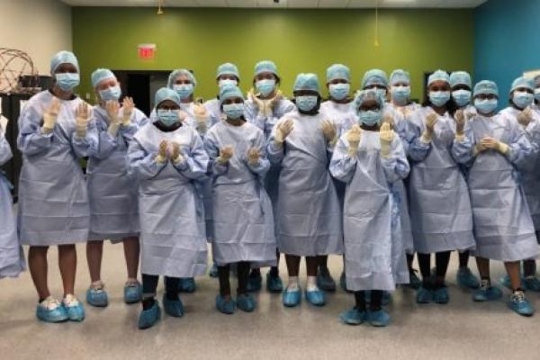 十九名高中生一起站在一间有绿色墙壁的房间里，穿着蓝色手术服、口罩、头发和鞋套。他们戴着手术手套，举起双手。