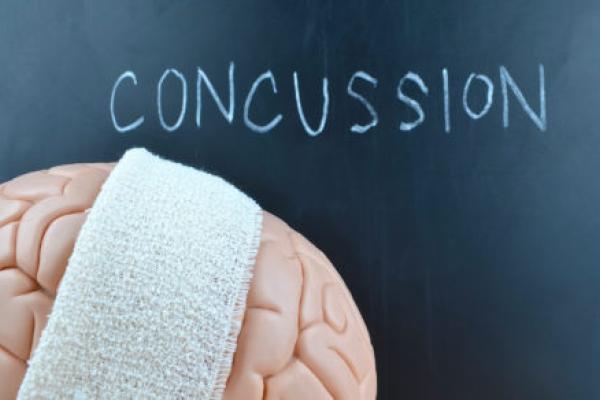 An illustration shows a brain draped with an athletic towel. Behind it on a chalkboard is written the word “concussion.”