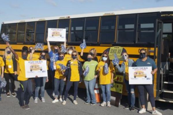 A group of 14 women stand in front of a school bus holding “Stuff the Bus” signs, pom-poms and foam fingers. Ten of them are wearing United Way T-shirts. Behind them is a Penn State Health pop-up tent.