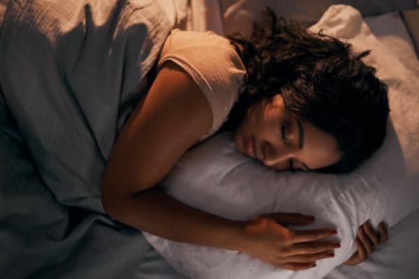 一个女人在左侧躺在床上，在一个被子下。她的眼睛闭着眼睛，她的手臂缠绕在枕头上。