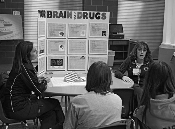 在“药物101”项目中，两名女护士坐在桌旁与两名青少年交谈。这些青少年背对着镜头。桌子上有一块三块的展示板，上面写着:“你的大脑和药物。”右边的护士正在说话。两位护士都穿着印有宾夕法尼亚州立大学标志的运动服。
