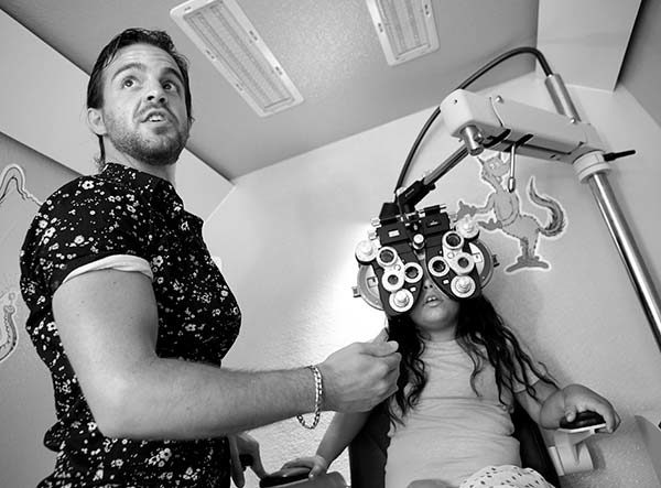 Aaron Harsch博士，一个透视学习的验光师，用一块带有许多镜片的设备测试Gabriella的景象，因为她坐在移动实验室的椅子上。Harsch穿着一件花卉实验室外套，留着胡子。