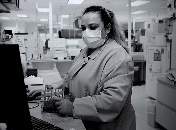 宾夕法尼亚州立大学医疗实验室的女科学家圣约瑟夫看着电脑屏幕，触摸着一盒试管。118金宝搏抽水她穿着磨砂服，戴着口罩，留着长长的马尾辫。她身后是实验室设备和灯光。