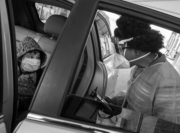 戴着面罩和听诊器的德西蕾·韦伯医生站在一辆汽车旁边，旁边站着一名戴着面罩和夹克的男孩。车门开着。