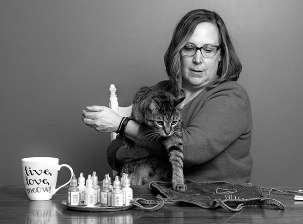 Wendy Johnson拥有肩长头发，佩戴眼镜，坐在桌子后面并握住她的猫，将一只爪子放在桌子上的牛仔布围裙上。瓶织物涂料和一个杯子说“活着，爱，喵”在桌子上。