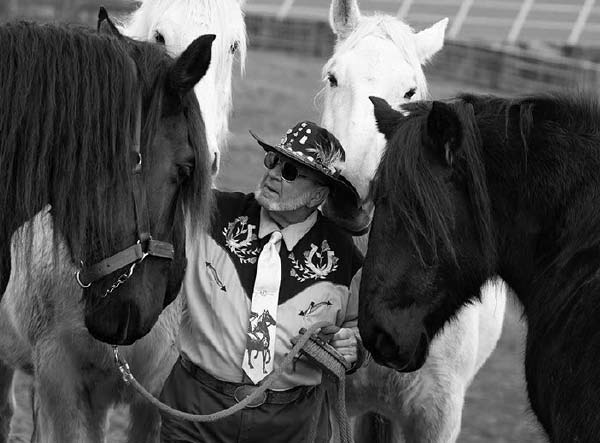 一个人站在四匹马中间，看着左边的那匹马。他留着胡子，戴着太阳镜，戴着一顶有羽毛的牛仔帽，穿着一件花背心，长袖衬衫和一条有马的领带。左边和右边的两匹马的前额耷拉在眼睛里。两匹白马的头在他身后。