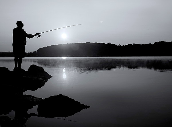 一个男人的剪影，面朝右，站在水边的一块岩石上，把鱼线投进湖里。太阳正落在一座被树木覆盖的小山后面，倒映在湖面上。
