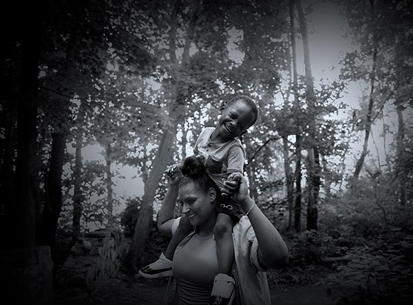 母亲笑着，儿子坐在肩膀上，笑着，握着母亲的手在树林里。