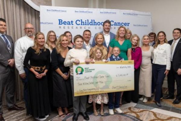 A group of adults and children pose with an oversized check in the amount of $3 million. A backdrop contains the Beat Childhood Cancer Research Consortium logo.