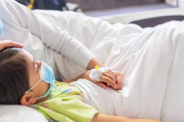 Young mother with protective face mask holding son's hand while lying on the hospital bed with him.