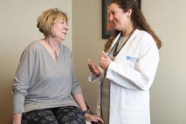 A woman sits on an exam table talking to Dr. Jaime Long, right, of Penn State Health. Dr. Long is gesturing with her hands and wearing a white coat with the Penn State Health Milton S. Hershey Medical Center logo on it. Both are smiling.