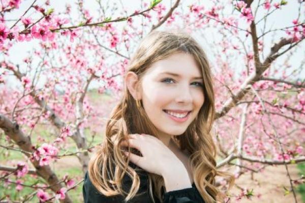 A teen girl in front of a flowering tree smiles at the camera. She has long hair and is touching her shoulder.