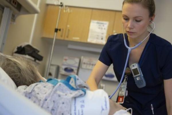 A nurse uses a stethoscope as she tends to a young patient in a hospital bed. The nurse also has a phone and a nametag clipped to her shirt. An IV pole and wooden cabinets are in the background.