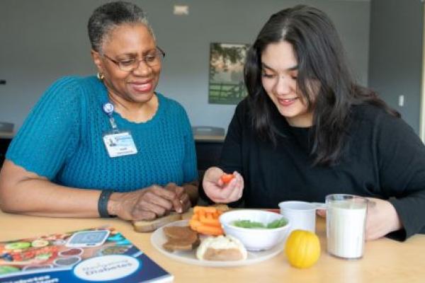 Two women sit at a table looking at a plate of food. A diabetes booklet is on the left. The woman on the left is wearing a knit top, glasses and a name badge. The woman on the right is wearing a long-sleeved top and is holding a cherry tomato.