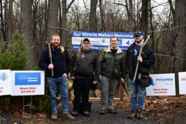 Four men pose for a photo at the edge of a wooded area. Three of them hold guns pointed at the ground.