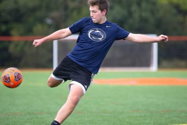 Brayden Sunho purses his lips as he gets ready to kick a soccer ball that is in mid-air. Brayden, who is wearing shorts and a shirt with a Penn State Nittany lion logo on the front, is running on a soccer field marked with goal lines.