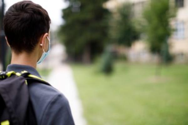 A boy wearing a mask and backpack stares down a sidewalk at a school building in the distance.
