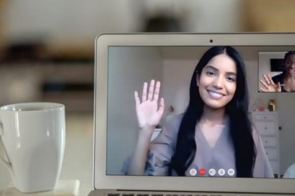 A woman in a video chat on a laptop screen waves to another, shown in a smaller window on the screen. A coffee cup is next to the laptop.