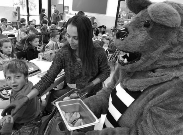 宾夕法尼亚州立大学PRO Wellness apple Crunch活动期间，一名妇女在拥挤的小学食堂分发苹果片。宾州州立大学的Nittany狮子吉祥物站在她旁边，手里拿着一桶苹果片。那个女人穿着一件毛衣，手上戴着塑料手套。一个小男孩看着镜头，脸上带着惊讶的表情。